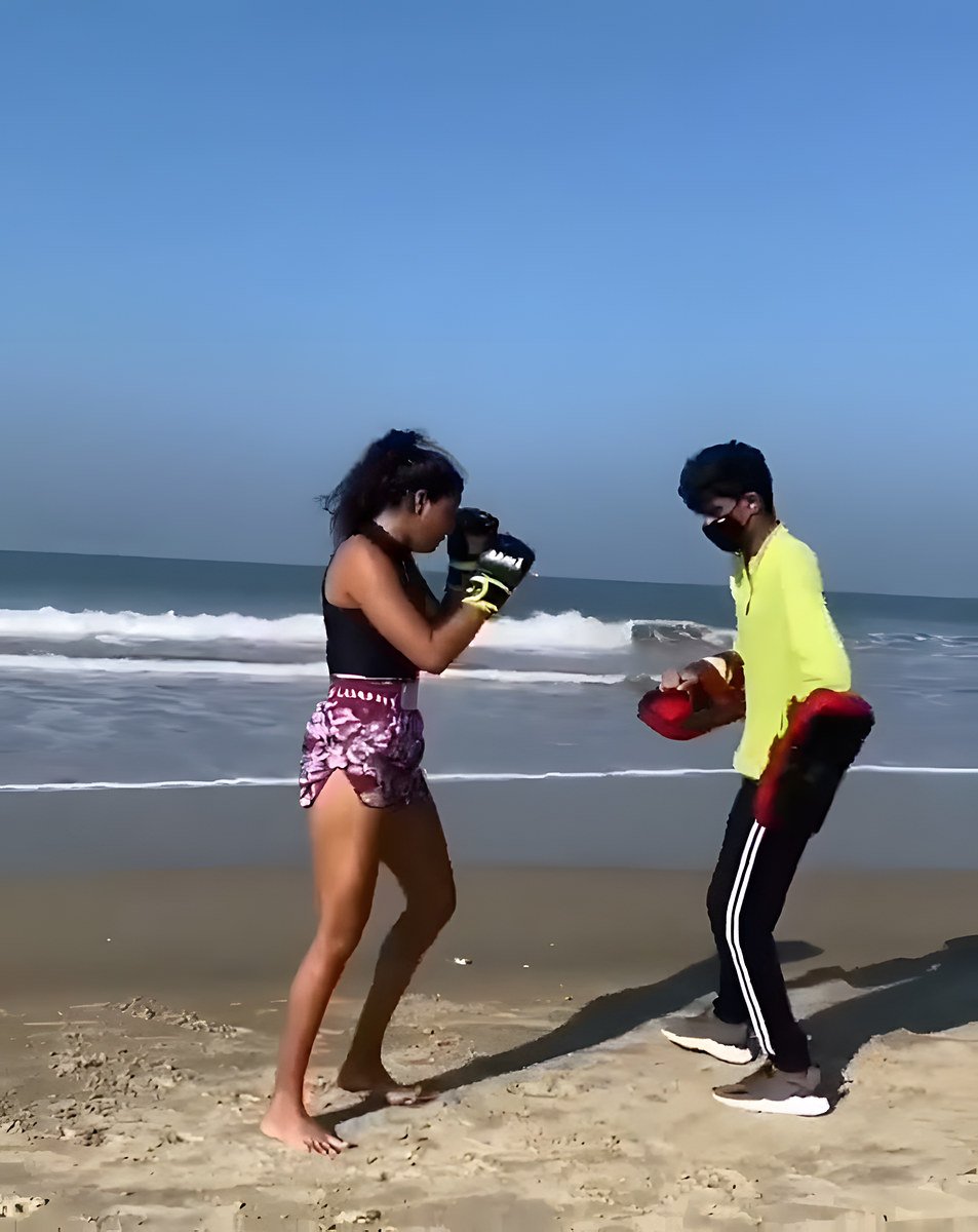 On a bright beach, a woman in a black top and pink Muay Thai shorts holds her guard while a masked man in a neon-yellow top holds pads.