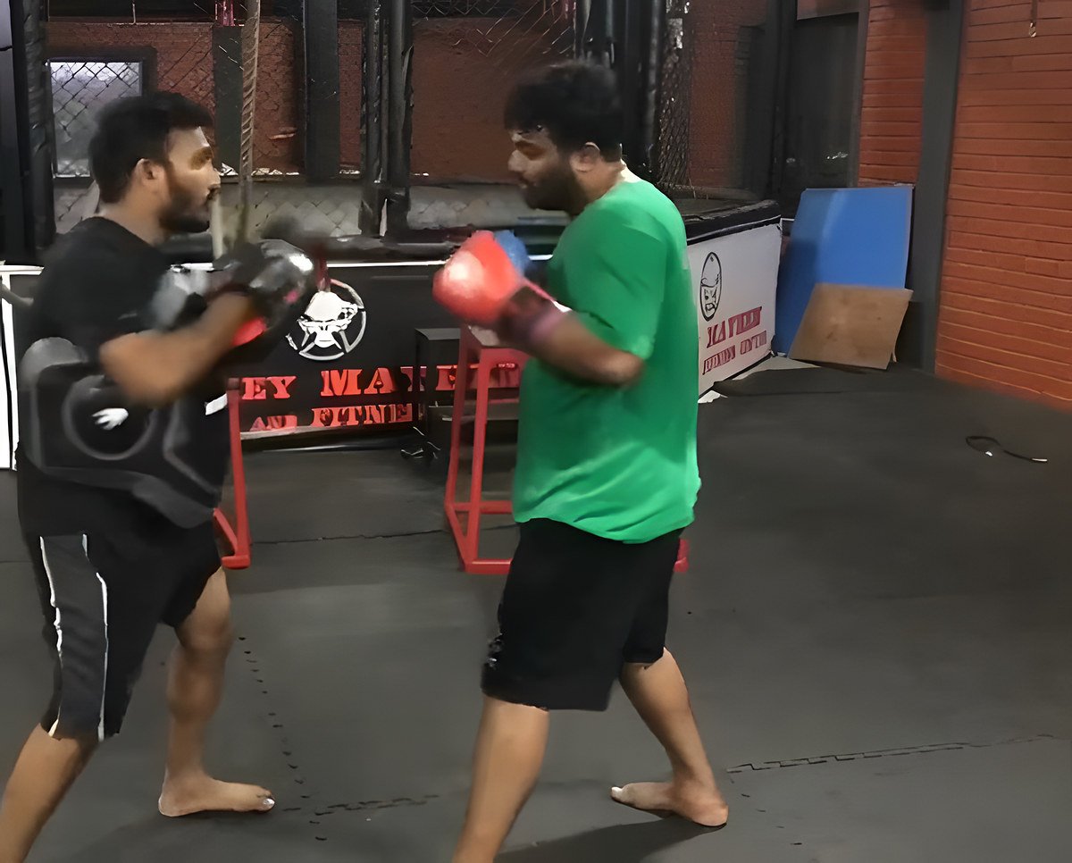 A bearded man in a green T-shirt and red gloves hits a belly pad held by a trainer in black, in front of a cage wrapped in a Monkey Mayhem banner.