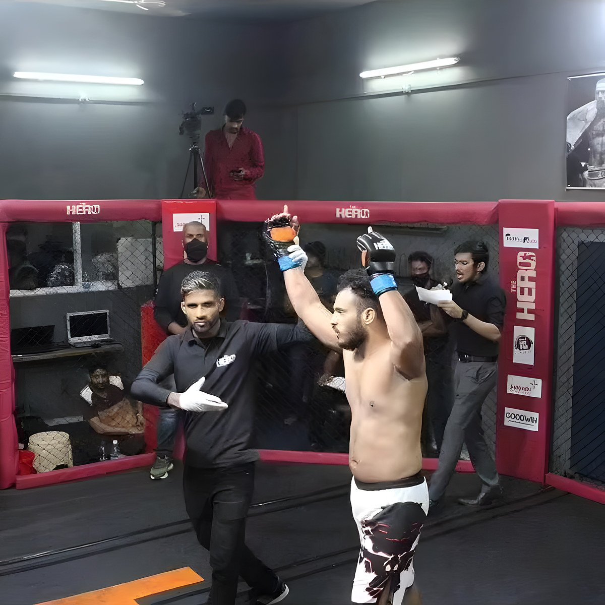 Inside a red-padded MMA cage, a shirtless fighter raises both gloved arms while an official in a black shirt gestures toward him.