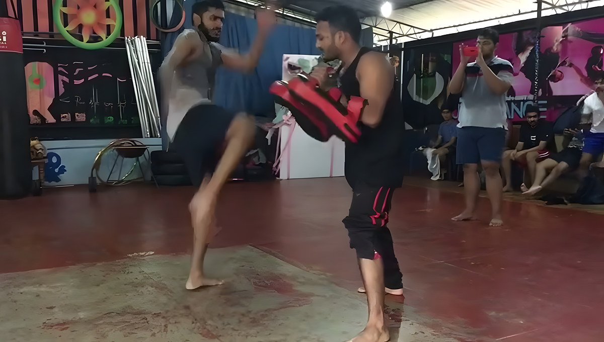 A man in a grey tank top kicks pads held by a man in a black tank top while others watch in a red-floor hall.