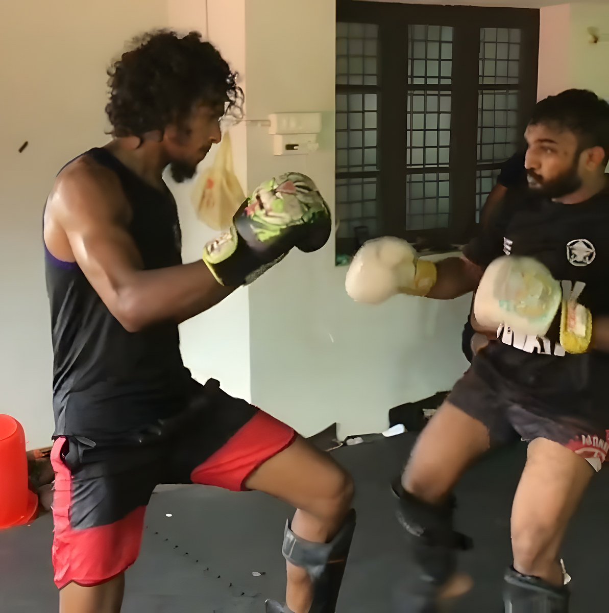 A curly-haired fighter in a black tank top and red shorts works with a bearded man in a black T-shirt holding pads, indoors.