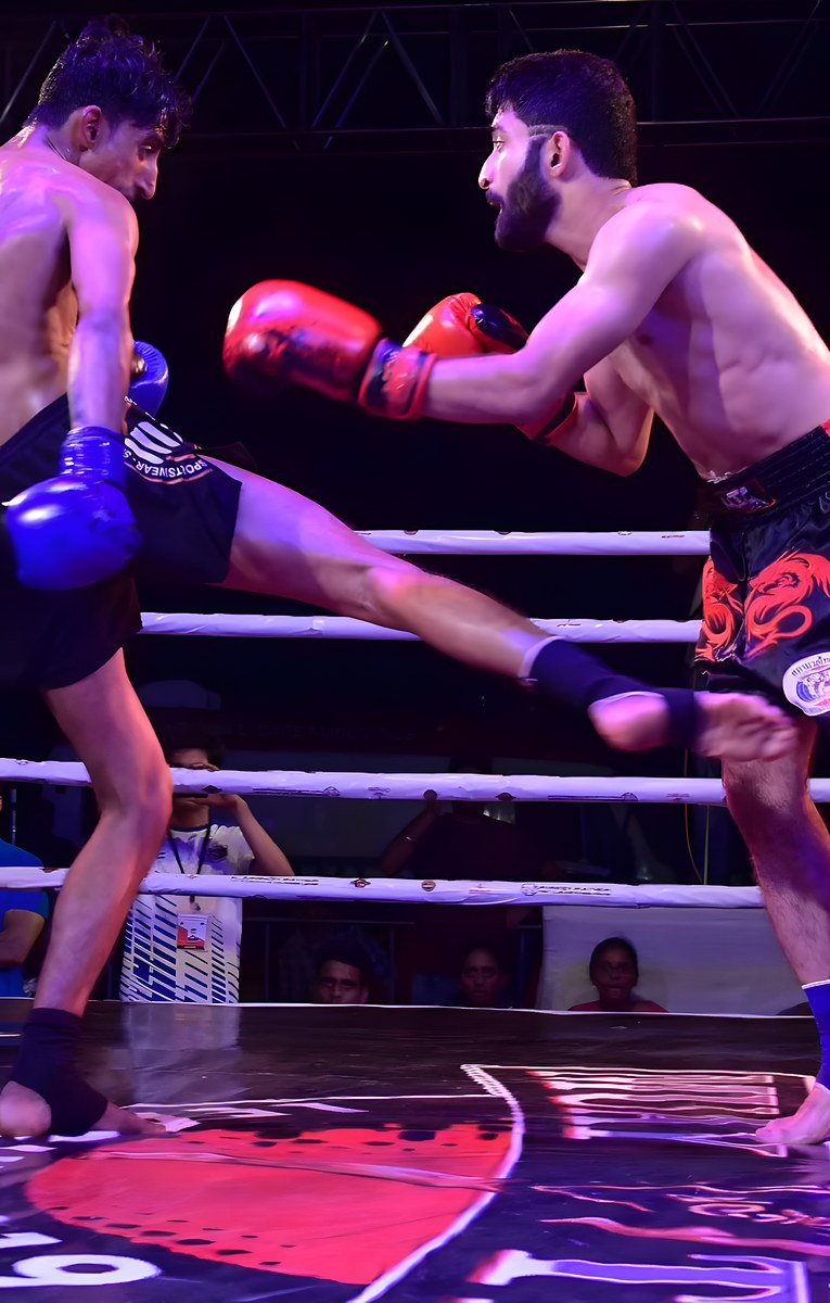 A shirtless fighter in black shorts and blue gloves throws a high kick at a bearded opponent in red gloves inside a ring with purple lighting.