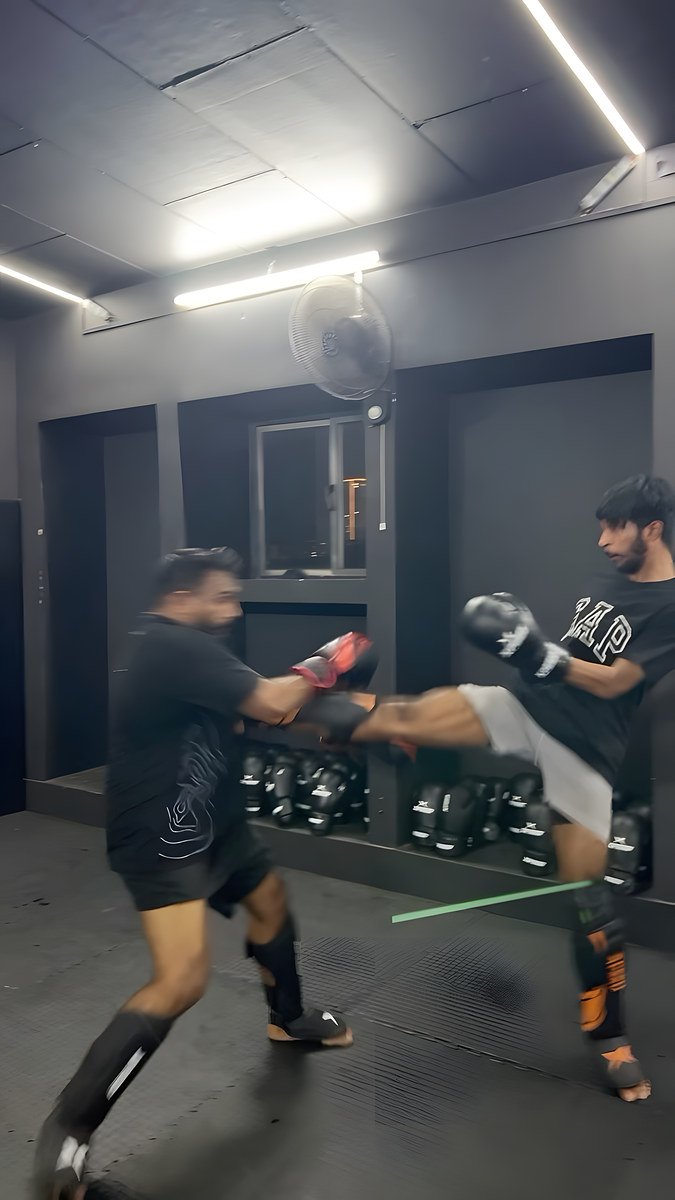 In the gym, a young man in a black T-shirt and shin guards throws a high round kick into the red pads of a bearded man in black.