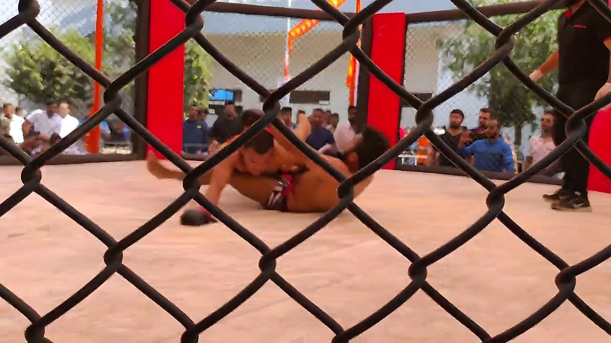 Two fighters grappling on the cage floor as a crowd watches through the fence