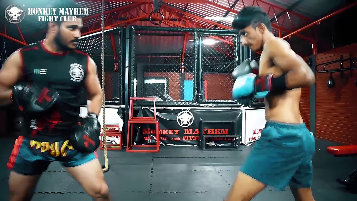 A trainer in a black-and-red Monkey Mayhem jersey holds focus mitts for a shirtless fighter in teal shorts and blue gloves, with a cage behind them.