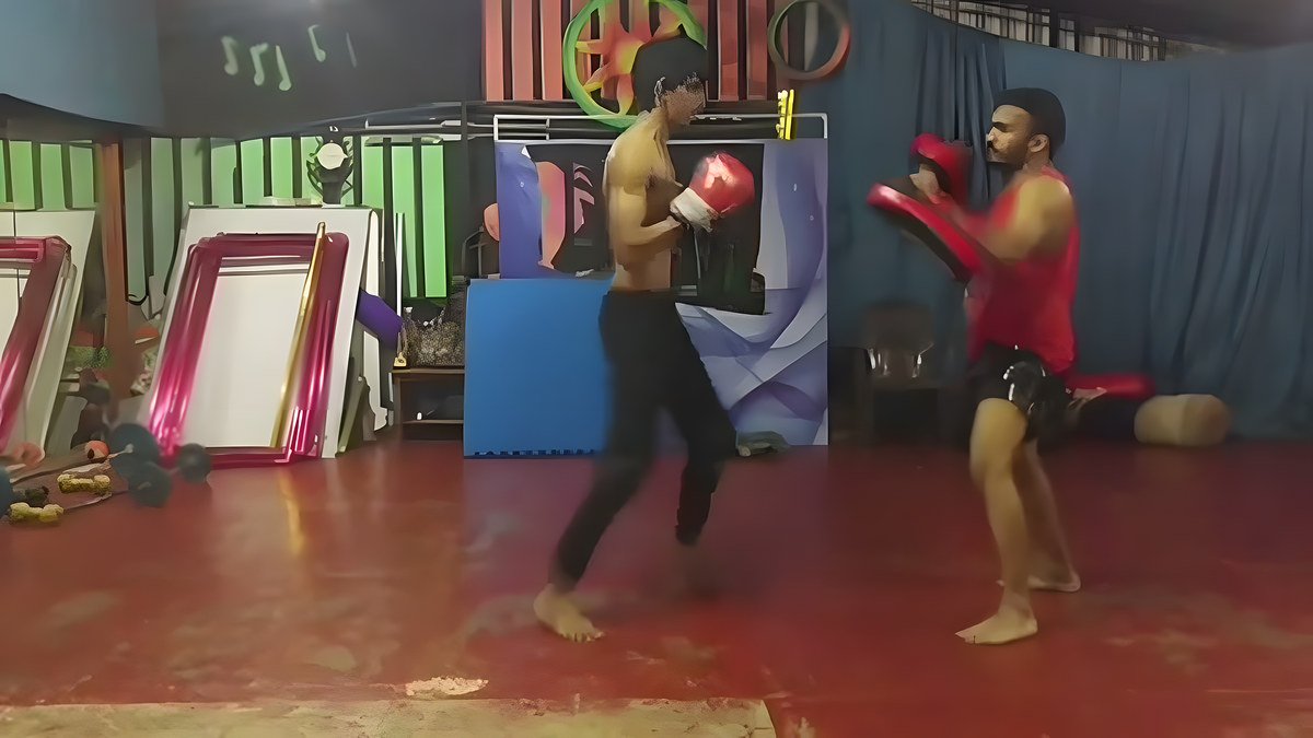 A shirtless young man in red gloves hits pads held by a man in a red tank top in a hall with a red floor and blue curtains.