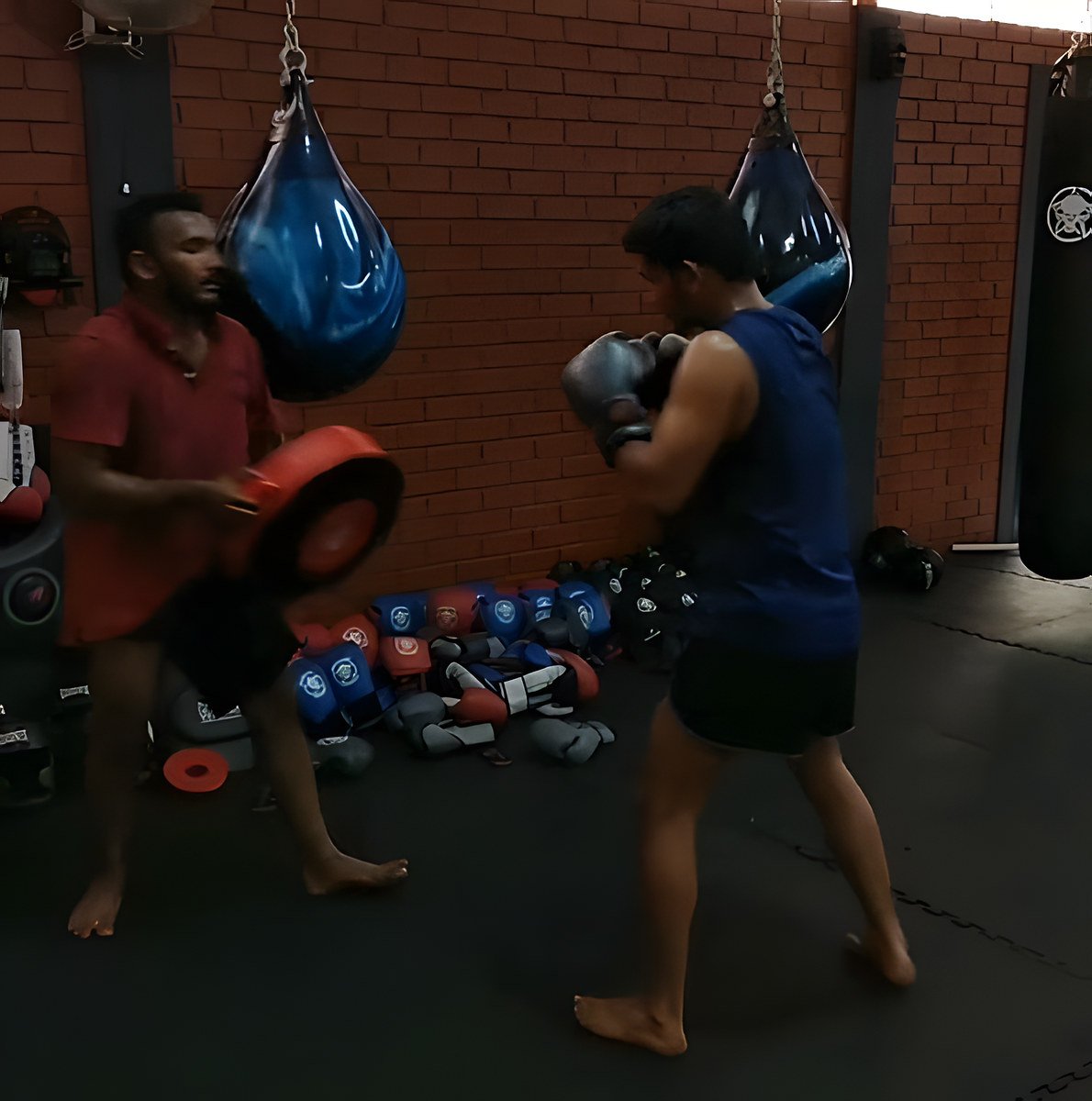 A trainer in a red shirt holds a punching shield while a fighter in a blue tank top throws, in the red-brick gym with aqua bags.