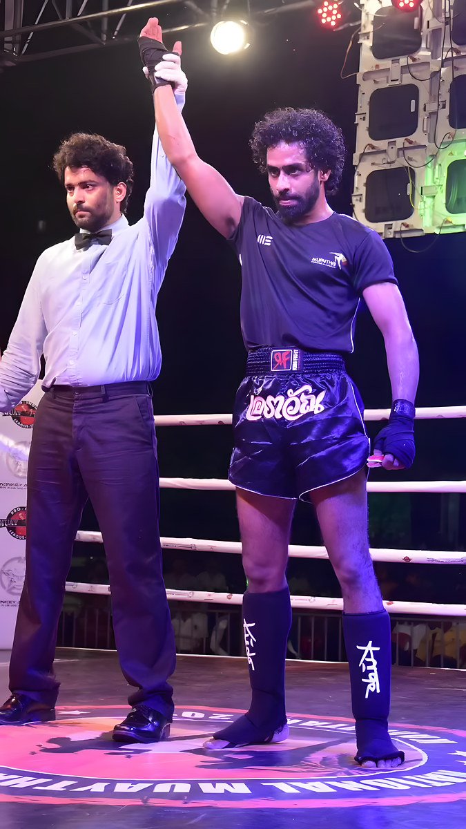 A referee in a bow tie raises the hand of a bearded fighter in a navy Muay Thai kit inside a lit ring.
