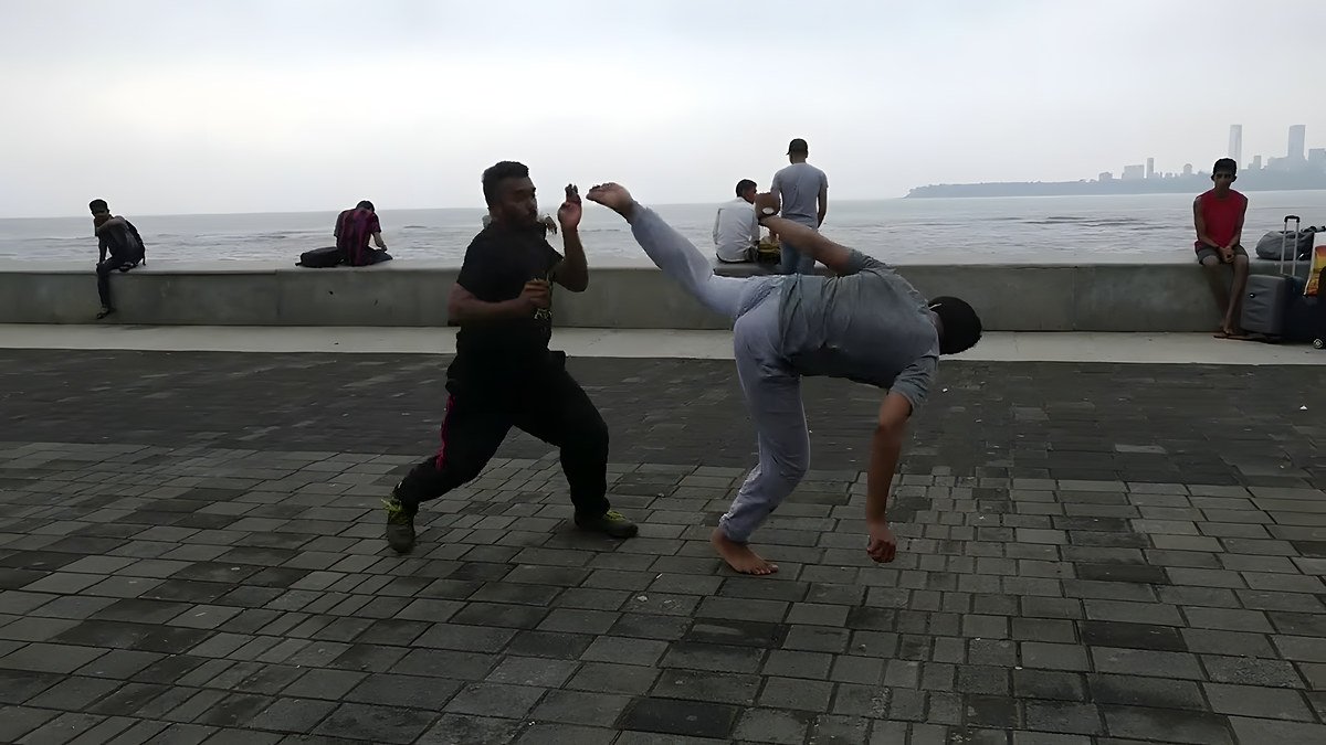 Two men on a seaside promenade with a city skyline behind; one in grey throws a low spinning kick, the other in black.
