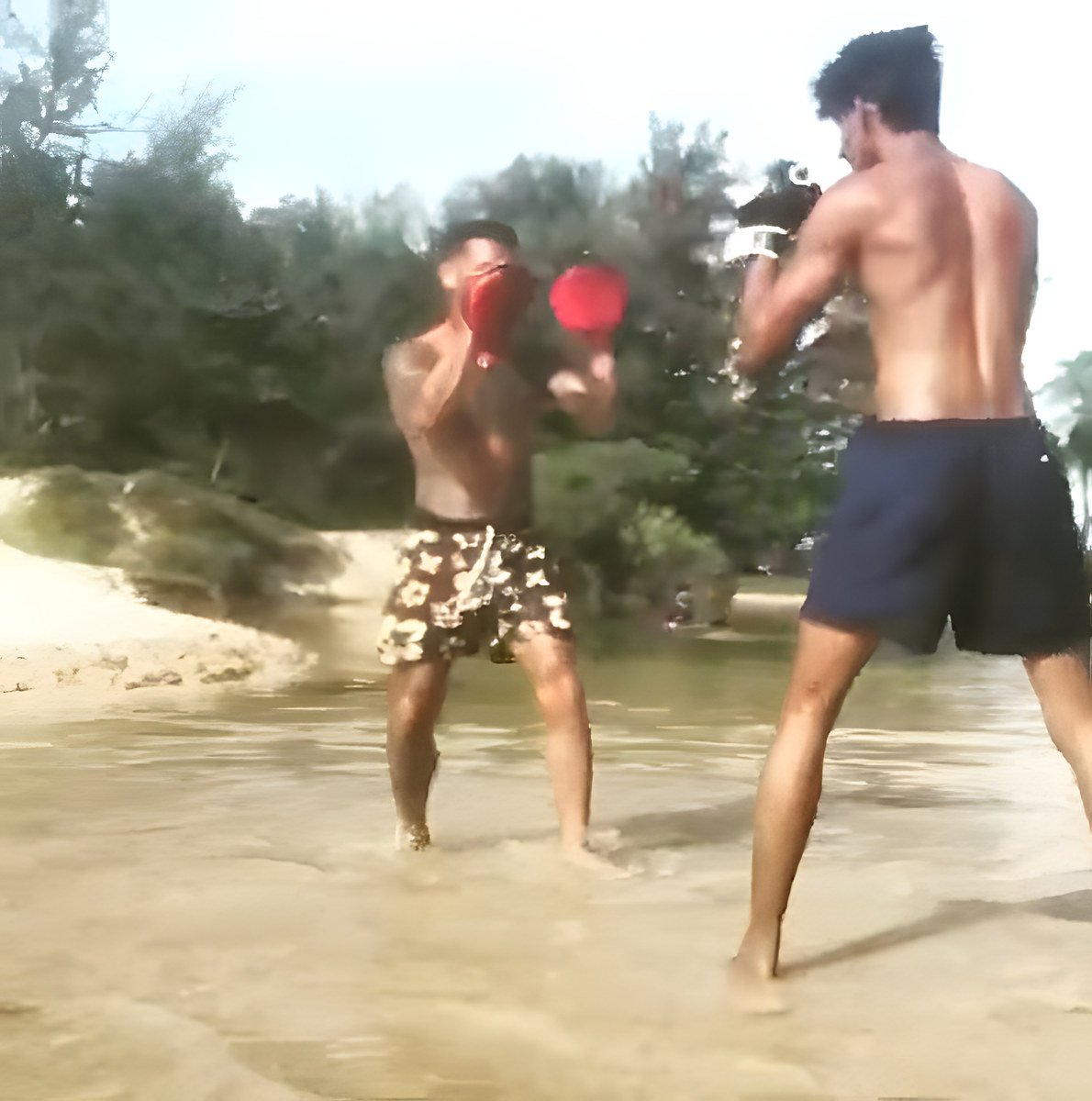 Two men train in shallow water; one in floral shorts holds red pads, the other is shirtless in navy shorts.