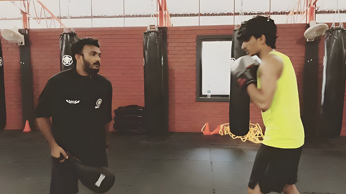 A bearded coach in a black club T-shirt holding a focus mitt works with a young fighter in a yellow vest in a gym with a brick wall and heavy bags.