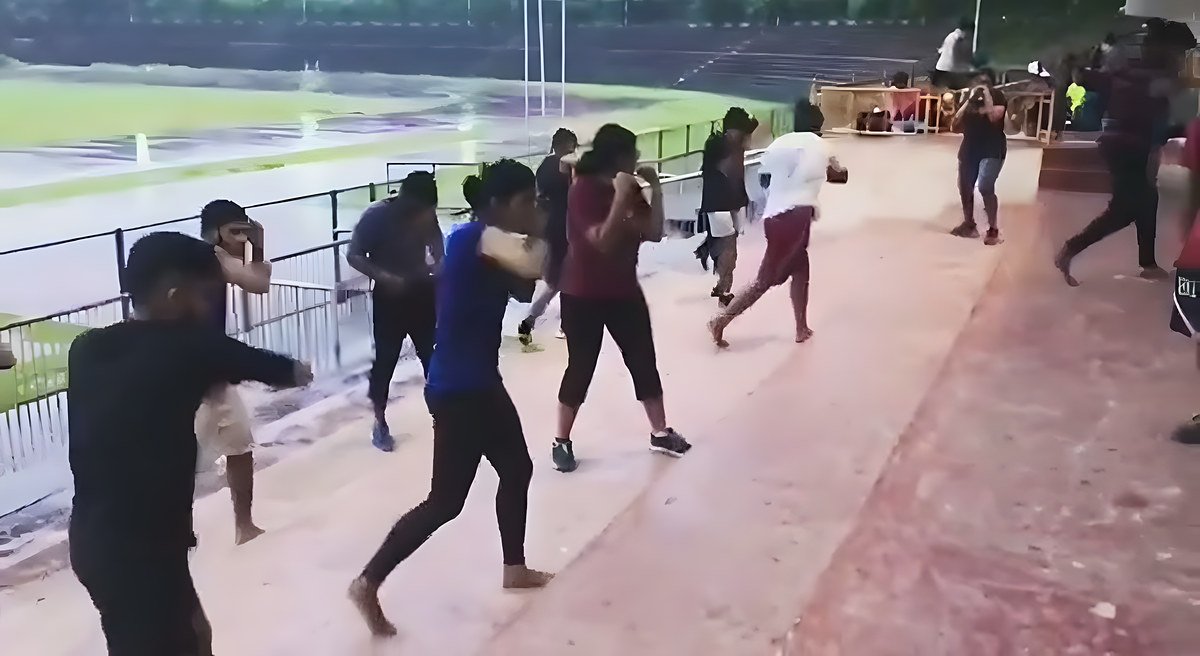 A group class practises elbows along a stadium stand walkway beside a rain-soaked track.