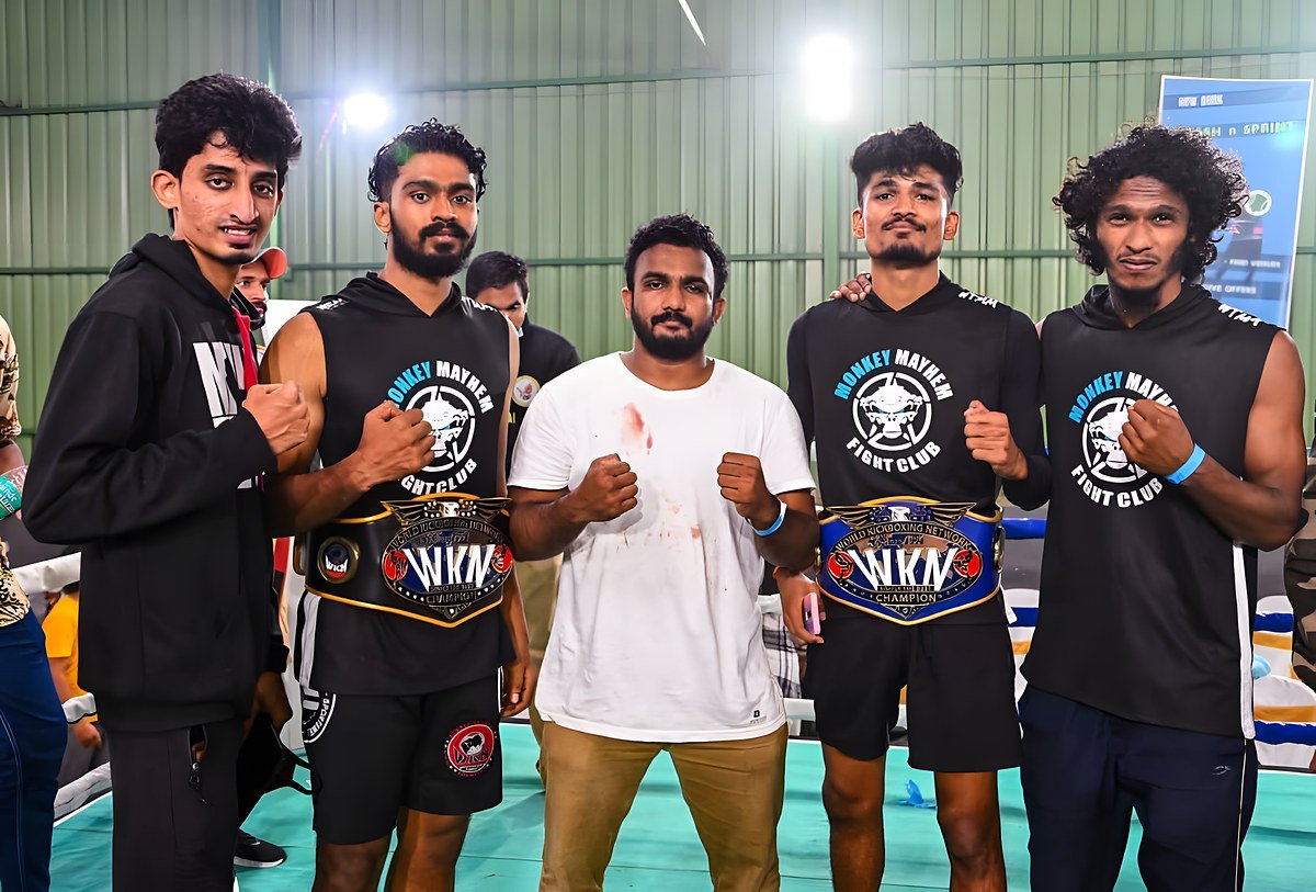 Five Monkey Mayhem team members in a ring, two wearing World Kickboxing Network champion belts, with coach Nithesh Kumar in the centre